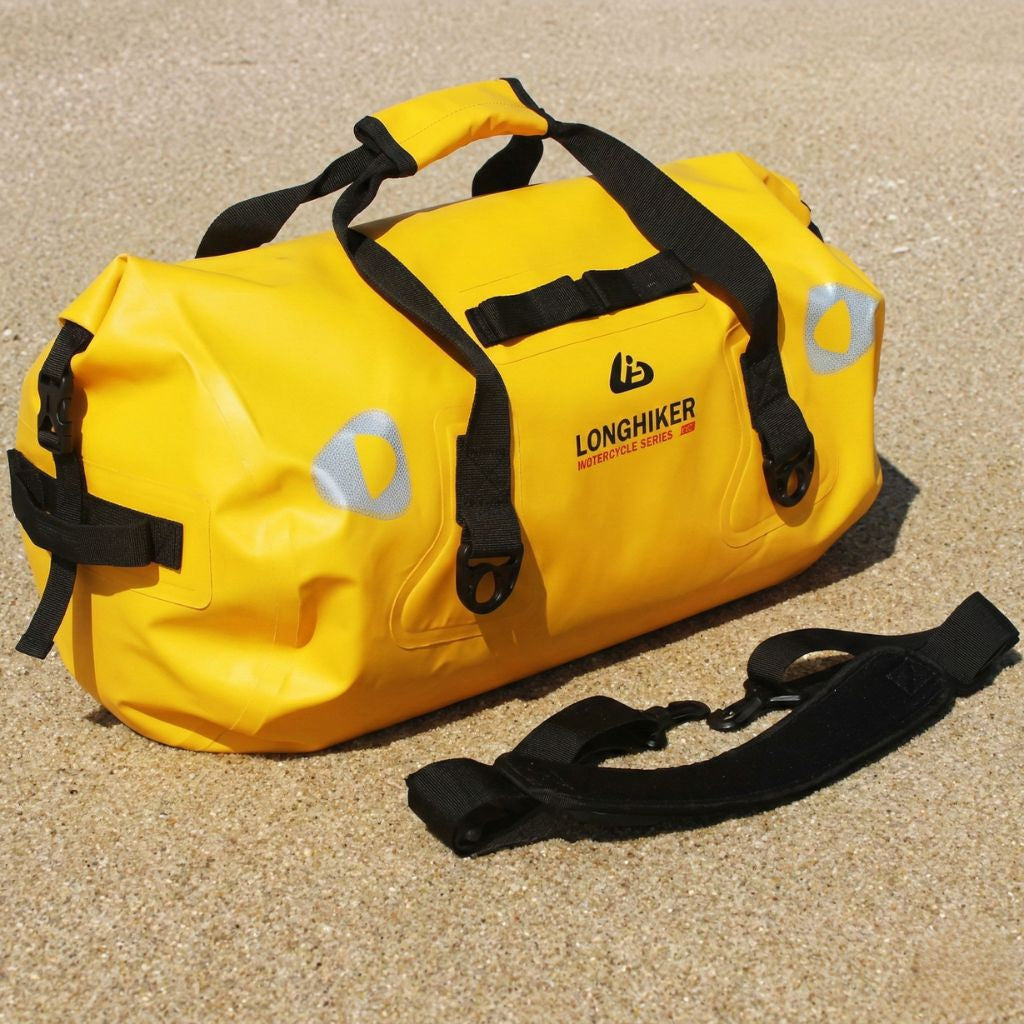 Sac de voyage étanche jaune 40L avec bandoulière posée à côté, vue 3/4 face sur fond de sable naturel