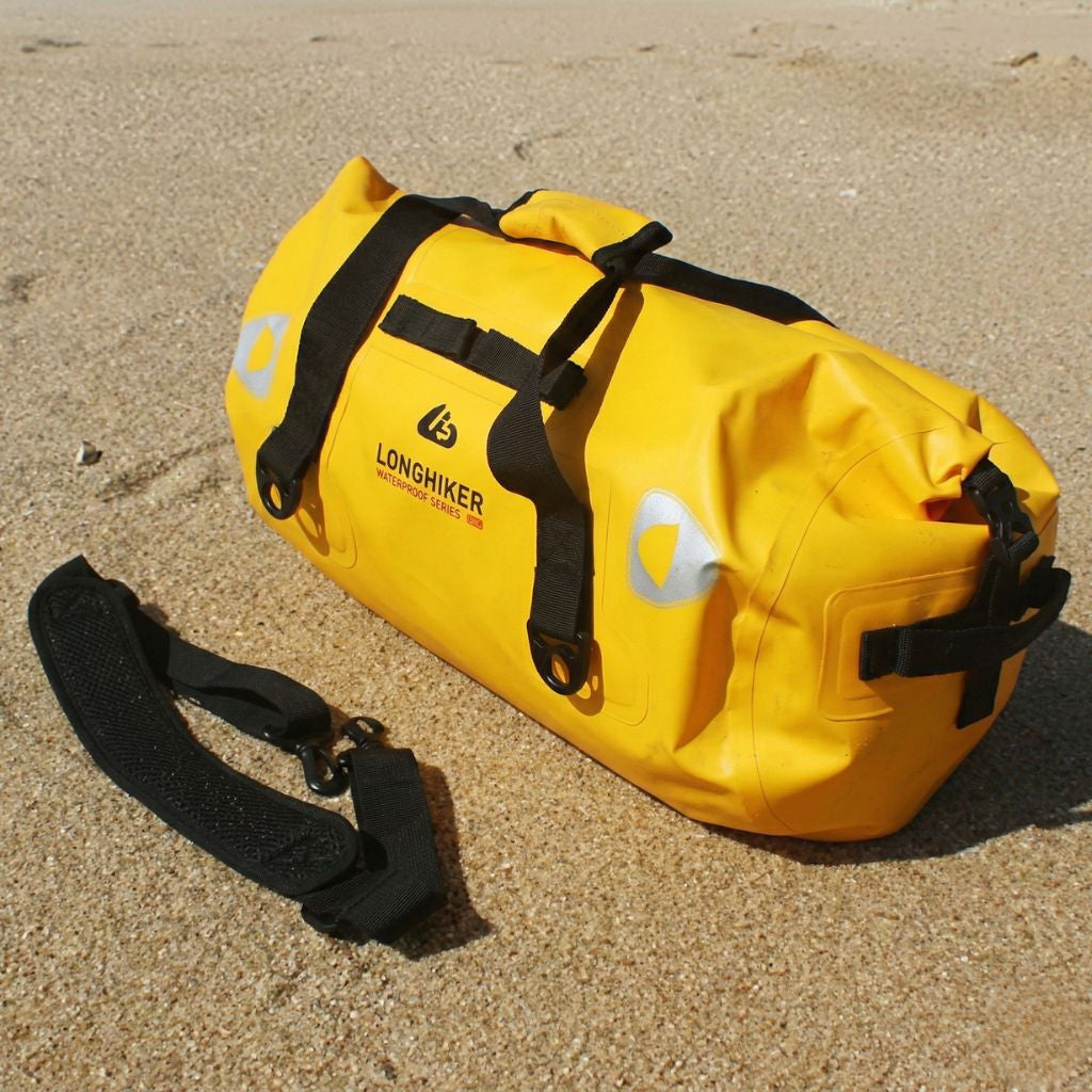 Sac de voyage étanche jaune format duffle posé sur plage de sable, vue 3/4, fermeture roll-top et sangles noires