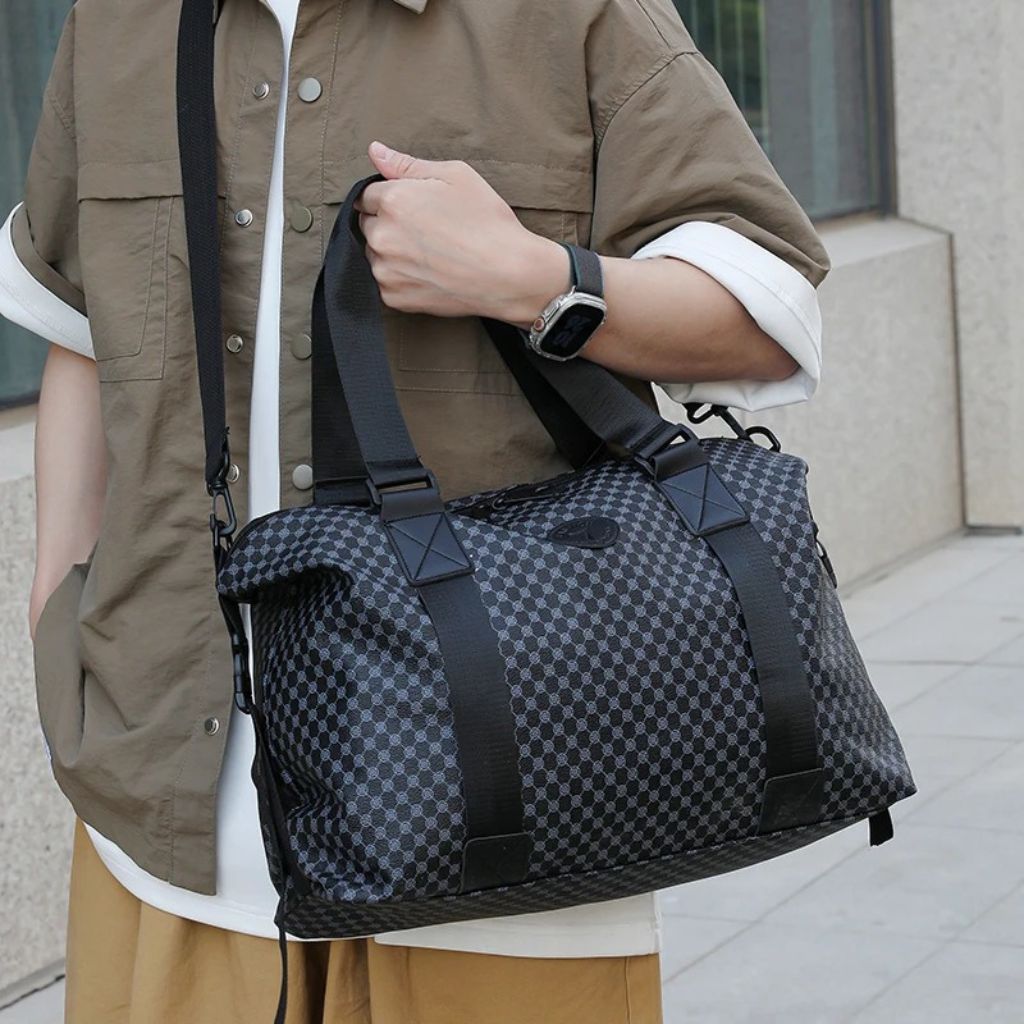 Homme portant le sac cabine damier gris anthracite en bandoulière et à la main dans un espace urbain