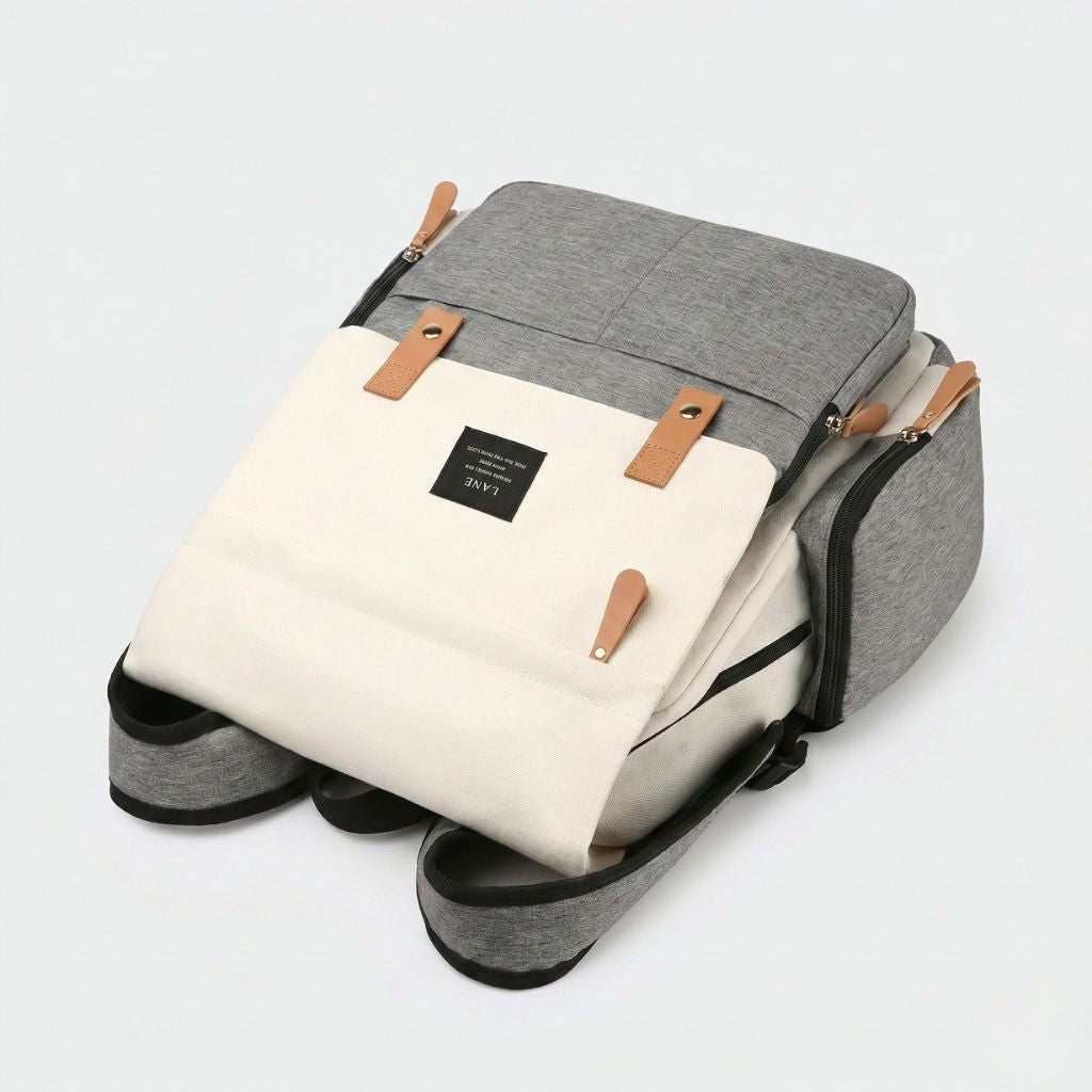 Sac à dos à langer bicolore gris et beige crème posé sur le côté, ouvert, avec le matelas à langer sorti