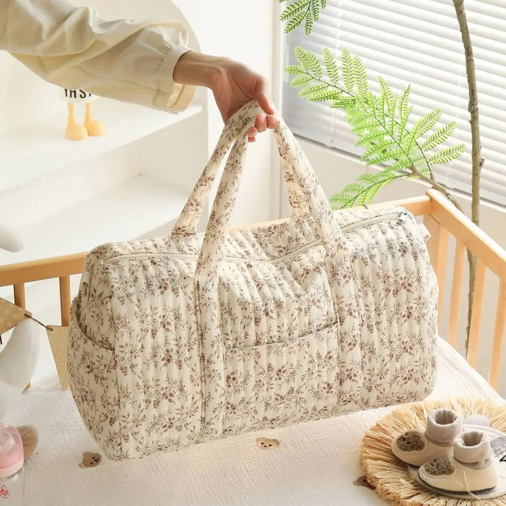 Sac à langer matelassé fleuri rose tenu par la poignée devant un lit de bébé, chaussons et biberon