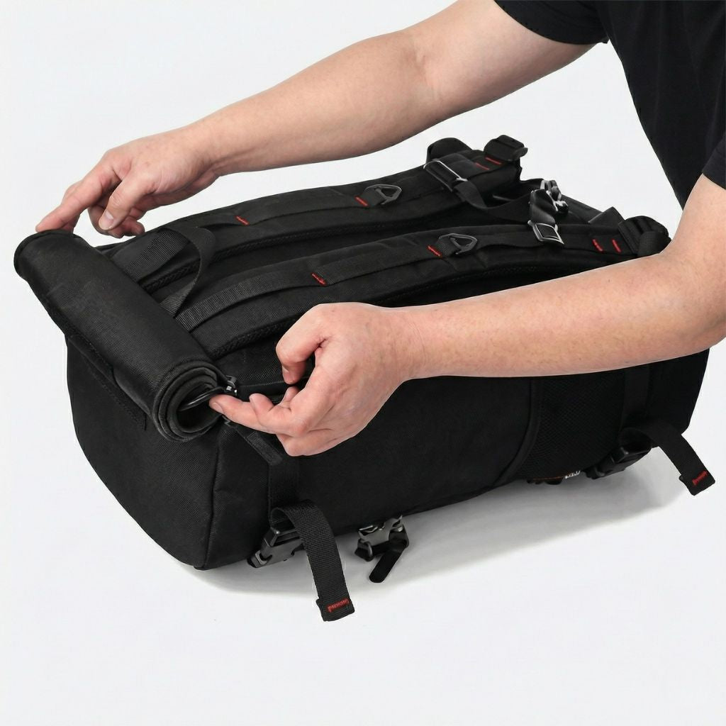 Grand sac noir tenu en vue plongeante, bretelles repliées et zip latéral d'accès principal bien visible