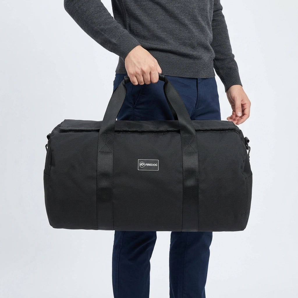 Homme en pull gris tenant un sac de sport cylindrique noir 34L par la poignée supérieure, pantalon bleu