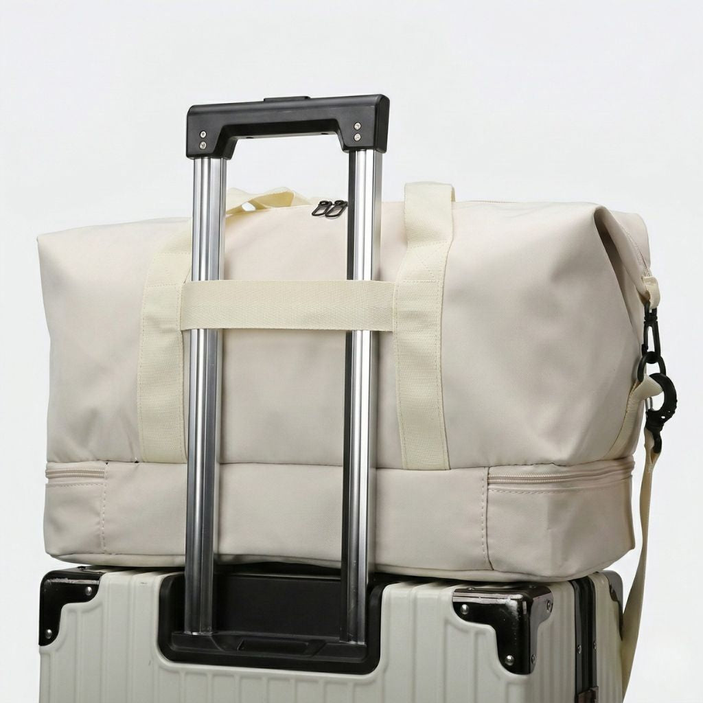 Sac de voyage de sport beige posé sur une valise blanche, sangle de maintien enfilée sur la poignée trolley
