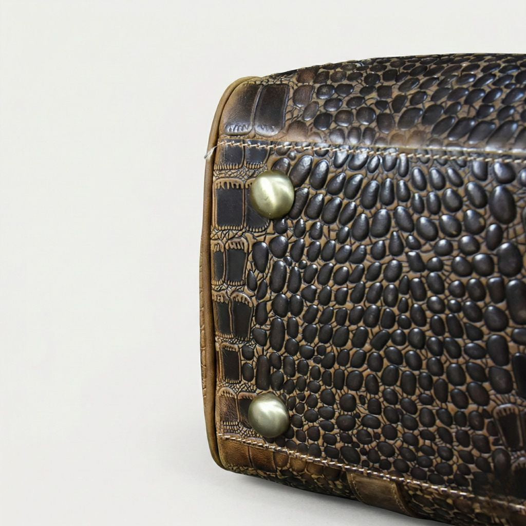 Gros plan coin du sac de voyage cuir croco brun, deux pieds protecteurs en laiton vieilli et motif reptile