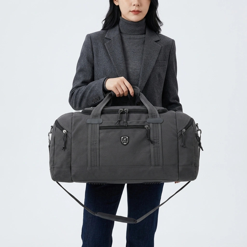 Femme en veste grise tenant un sac de voyage gris foncé par les poignées devant elle, port main