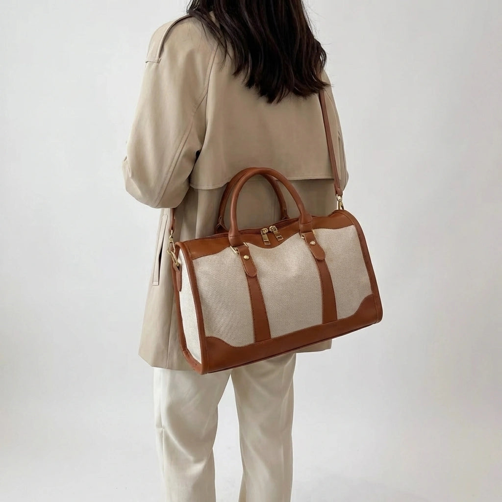Sac de voyage en toile beige et cuir cognac porté en bandoulière sur l'épaule, mannequin de dos en trench