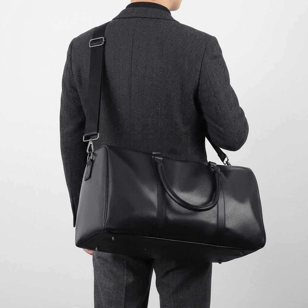 Homme de dos portant un sac de voyage en simili cuir noir en bandoulière, veste grise et pantalon