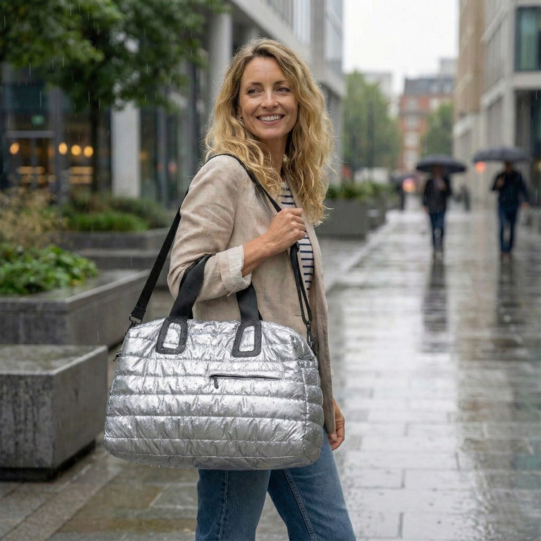 Sac de voyage matelassé grand format souple argenté avec anses noires et bandoulière amovible porté par une femme en ville sous la pluie Image