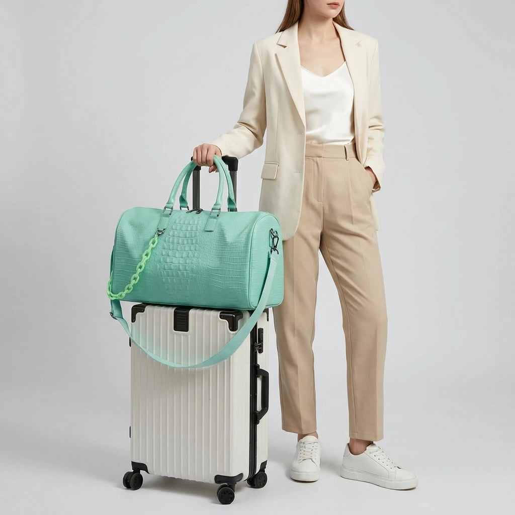 Sac de voyage vert menthe croco posé sur une valise blanche, mannequin en blazer beige, système trolley Image