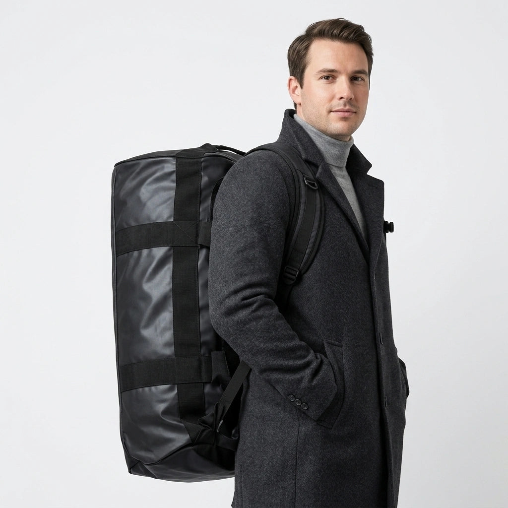 Homme en manteau gris portant un sac de voyage duffel noir 90L en mode sac à dos, bretelles réglables visibles