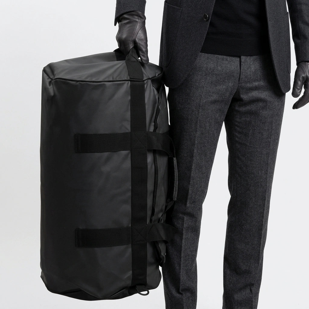 Homme en costume gris tenant par la poignée un grand sac de voyage duffel noir 90L, port à la main