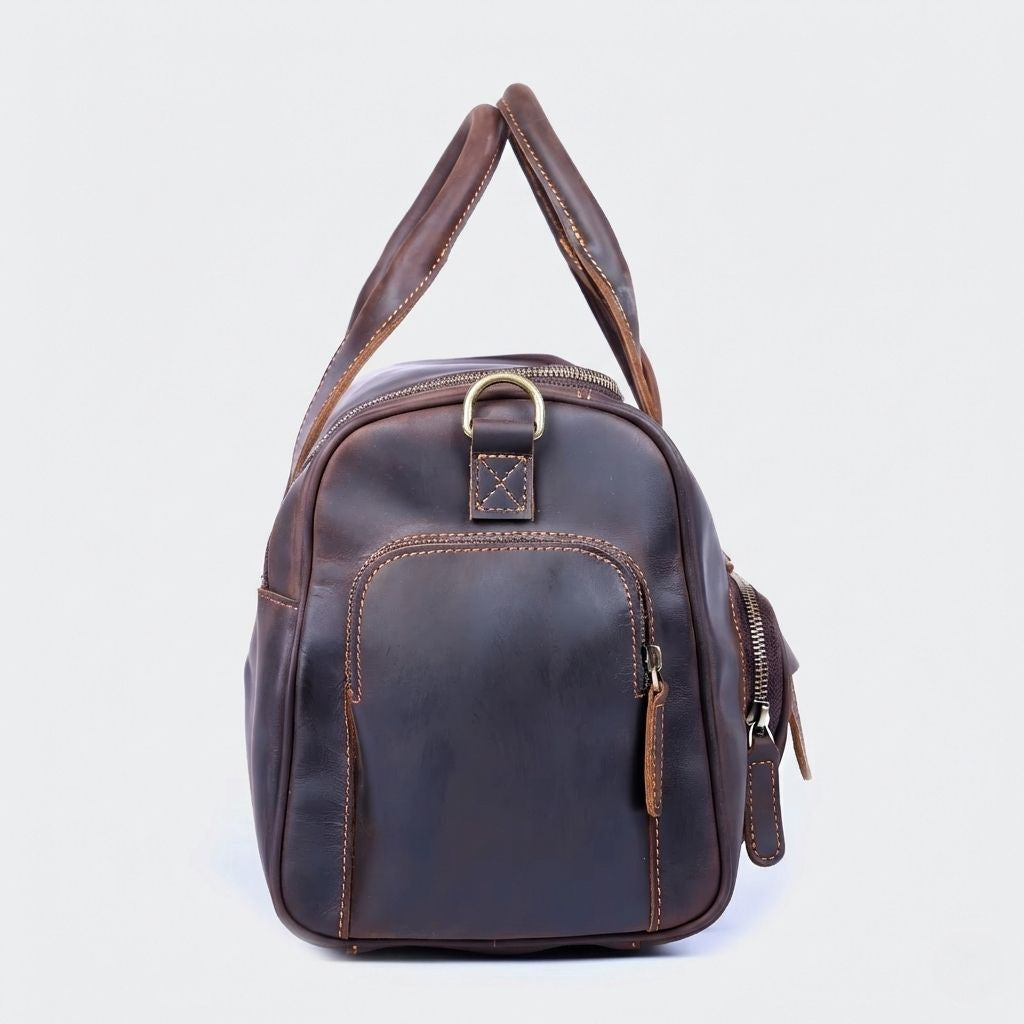 Sac de voyage en cuir marron foncé vu de profil, poignée arrondie, anneau laiton et poche latérale zippée