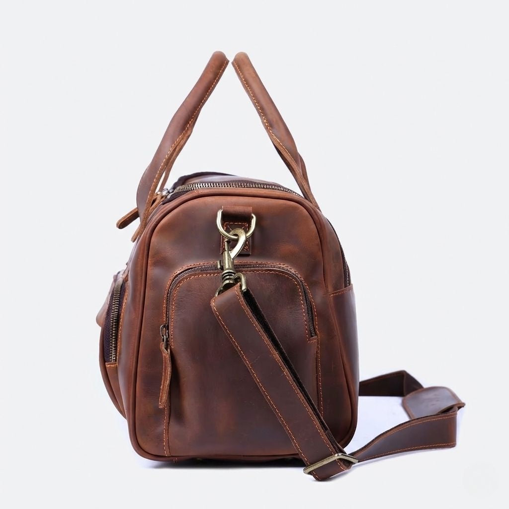 Sac de voyage en cuir marron cognac vu de profil, poignées rondes, fermoir laiton doré et poche latérale zippée