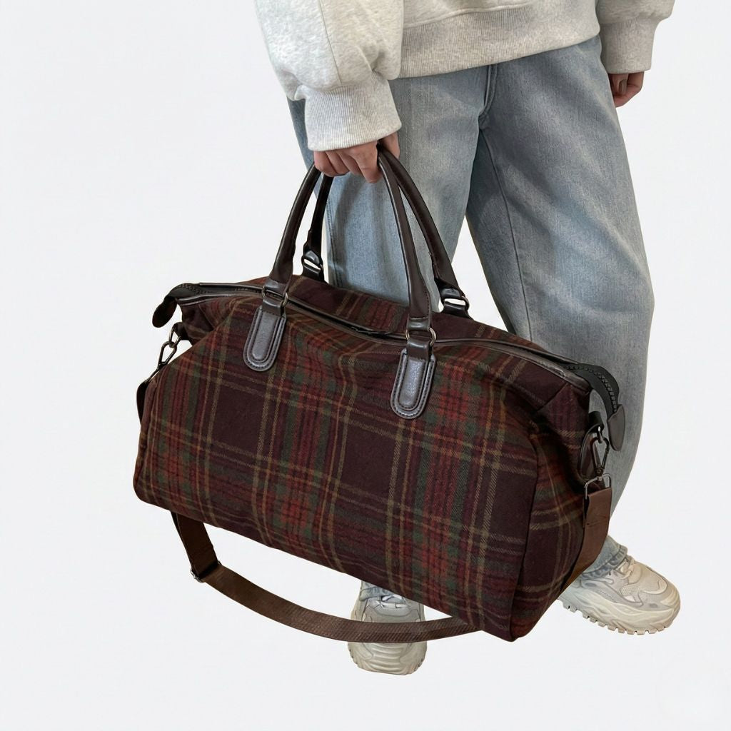 Personne tenant à la main un sac de voyage en toile tartan bordeaux, poignées cuir brun renforcées, style vintage