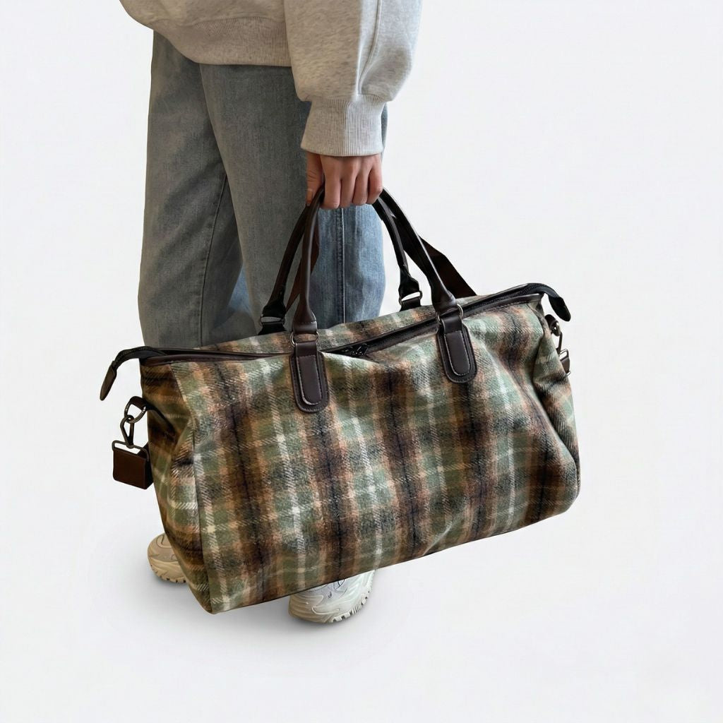 Personne tenant à la main un sac de voyage en toile tartan kaki, poignées cuir brun, style décontracté vintage