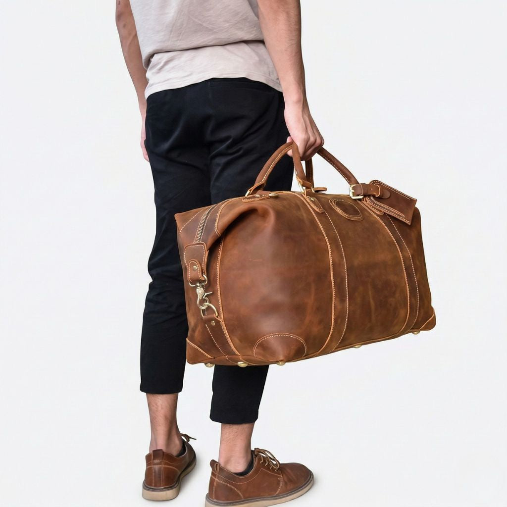 Homme portant le sac de voyage en cuir cognac par les poignées, côté arrondi et pieds laiton visibles Image
