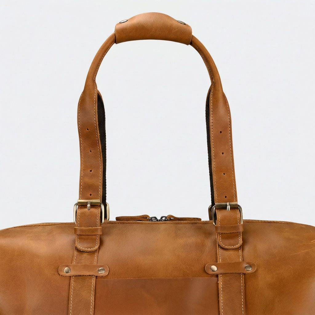 Détail des deux larges sangles de portage cuir cognac, boucles laiton et poignée rembourrée vue de face