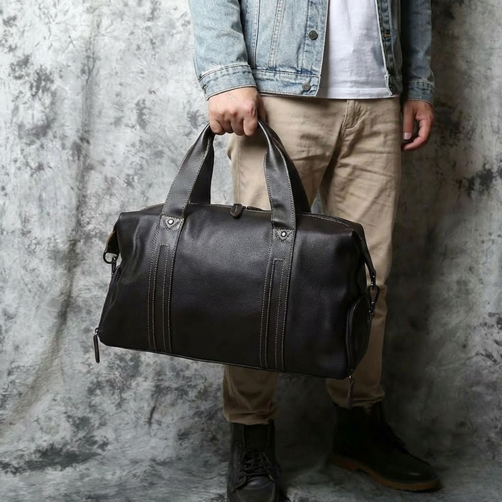 Homme tenant un sac de voyage en cuir noir par ses poignées, vue en pied devant un fond texturé marbré gris Image