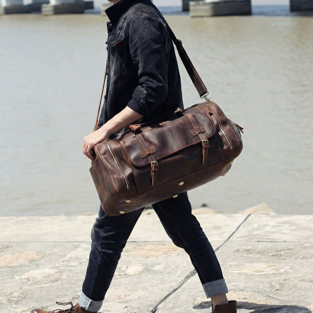 Homme marchant au bord de l'eau avec le grand sac de voyage en cuir brun chocolat en bandoulière