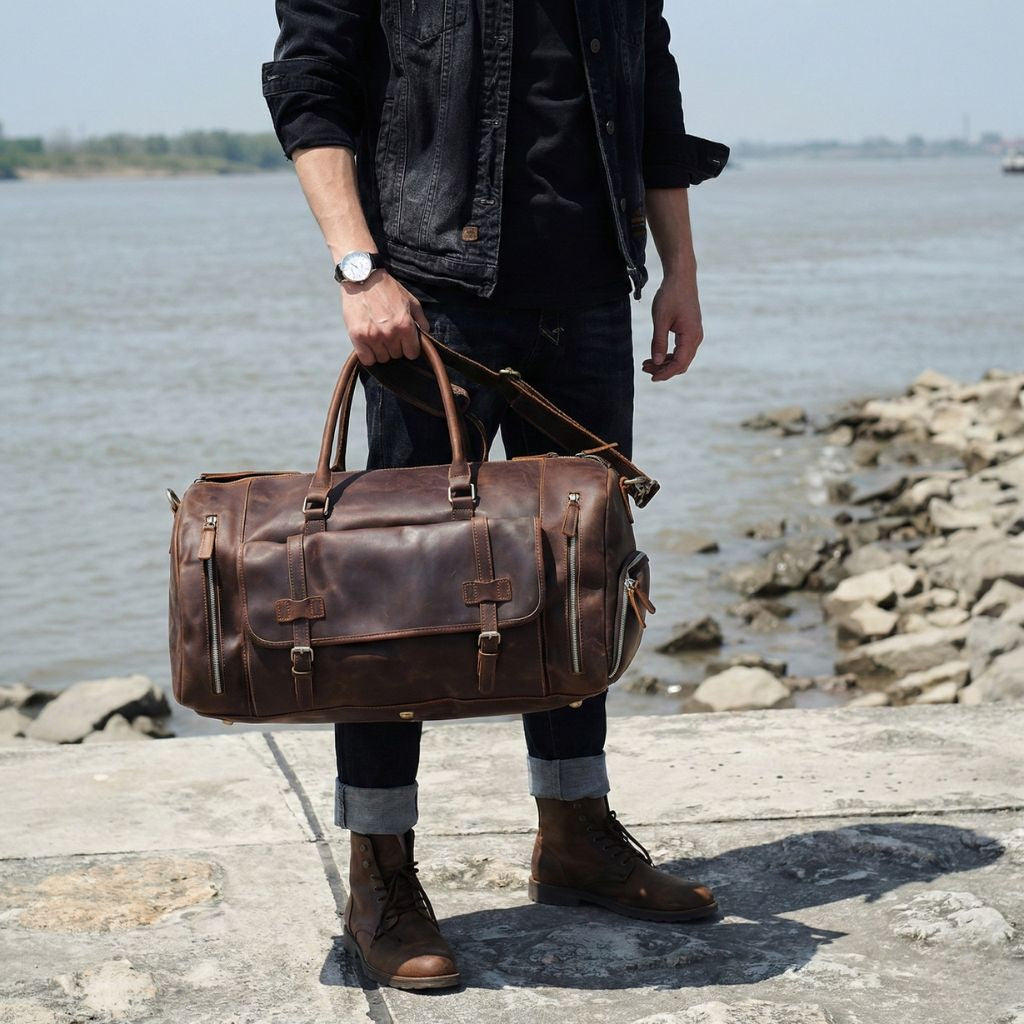 Homme debout en bord de mer tenant le sac de voyage en cuir brun par les poignées, look urbain