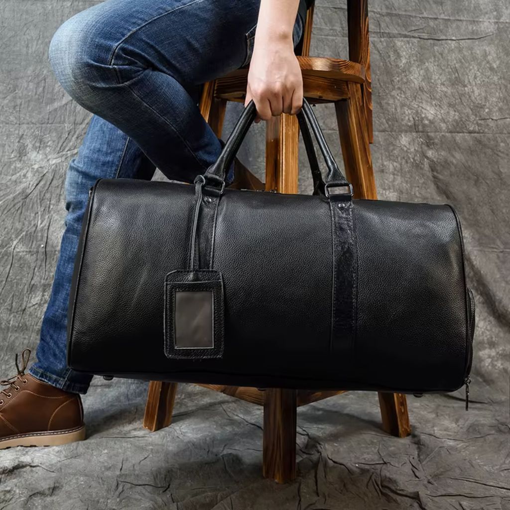 Sac de voyage cuir grainé noir porté à la main par un homme en jeans sur un escalier en bois