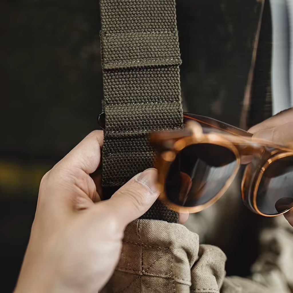 Détail de la sangle en toile kaki olive du sac de voyage militaire, mains tenant la bandoulière et lunettes écaille