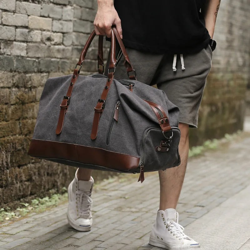 Homme portant à la main un grand sac de voyage en canvas gris anthracite et cuir marron, en extérieur Image