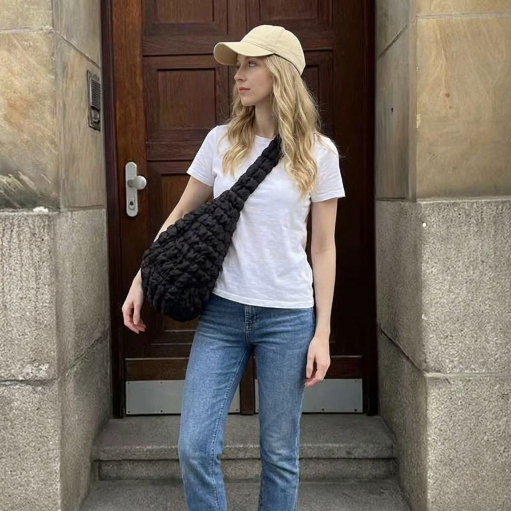 Femme portant un sac de voyage bandoulière matelassé noir devant une porte en bois, tenue jean et t-shirt blanc Image