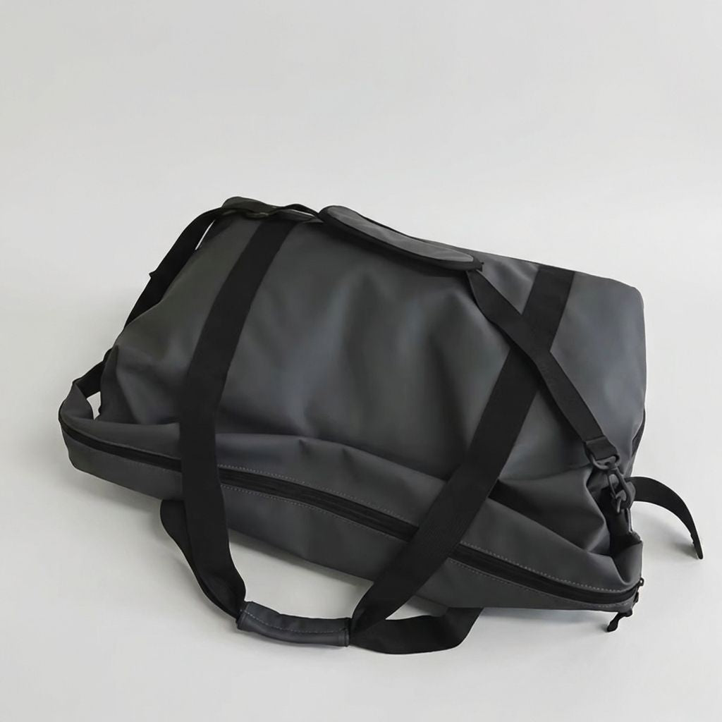 Sac de voyage imperméable noir graphite posé à plat : sangles noires croisées et poche arrière zippée visible