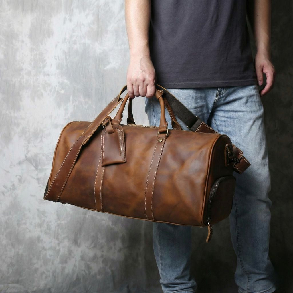Homme en jean et t-shirt gris tenant un sac de voyage en cuir cognac par les poignées, fond béton texturé