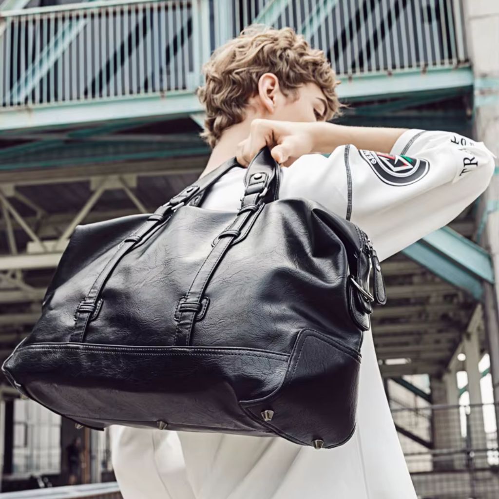 Jeune homme portant un grand sac de voyage en cuir PU noir en bandoulière sur l'épaule, milieu urbain industriel Image