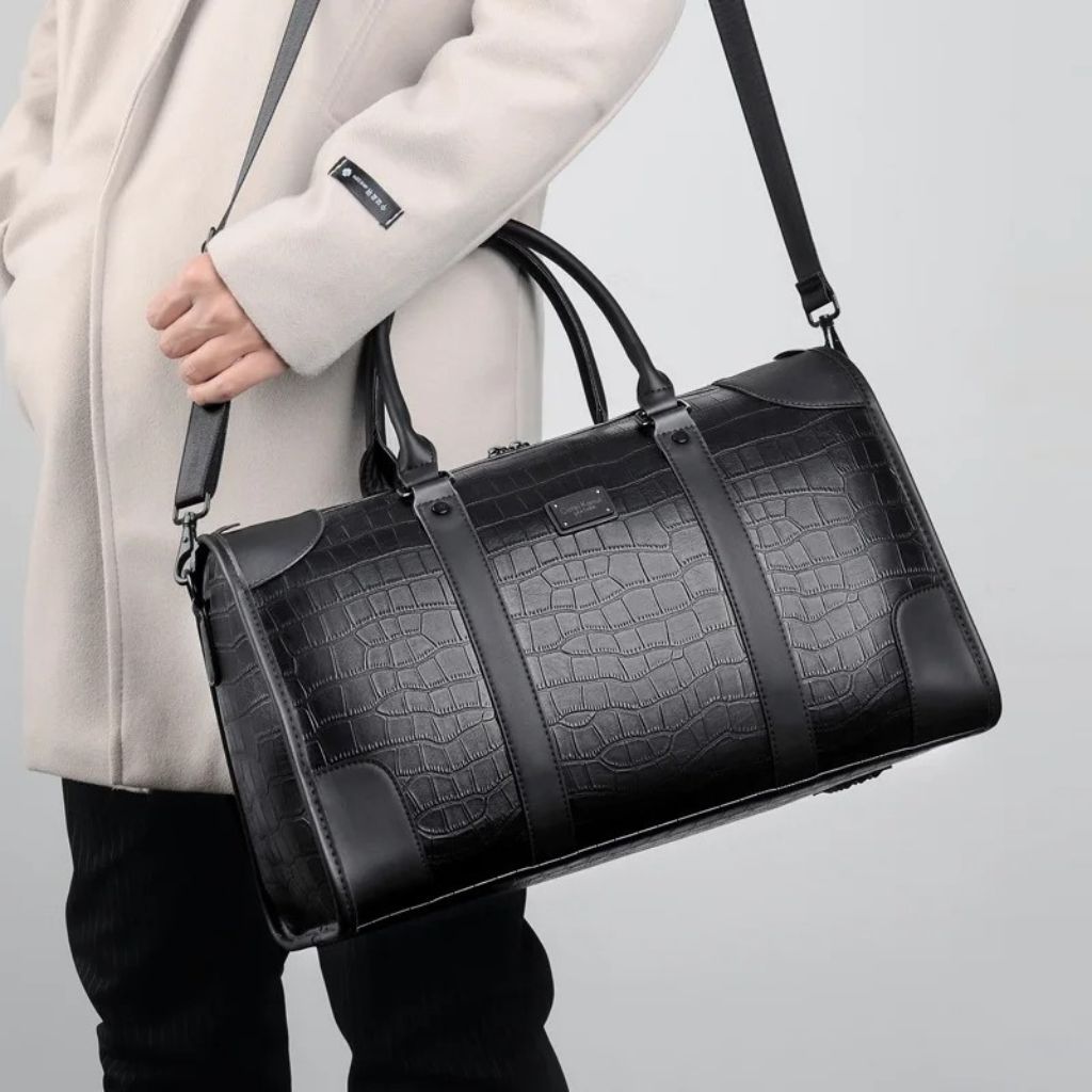 Homme en manteau beige tenant d'une main un sac de voyage en cuir noir aspect croco, bandoulière à l'épaule Image