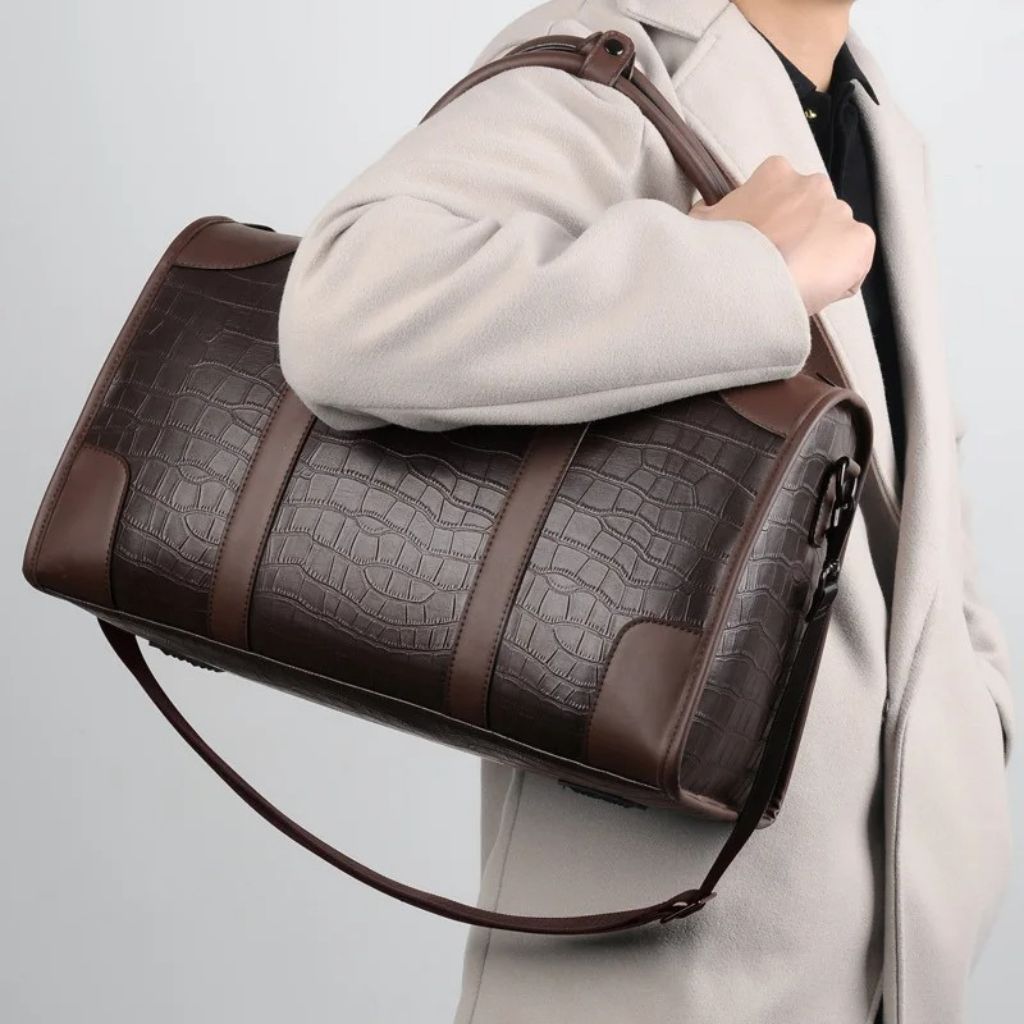 Homme en manteau beige portant en bandoulière un sac de voyage en cuir brun aspect croco compact Image